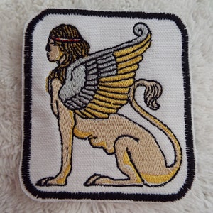May include: Embroidered patch featuring a white background with a gold and silver winged lion with a human head. The patch has a black border.