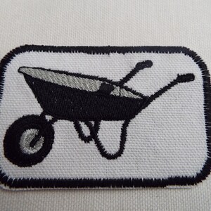 May include: A black and white embroidered patch of a wheelbarrow. The patch has a black border and is on a white background.