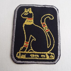 May include: A black embroidered patch with a gold outline of an Egyptian cat sitting on a platform. The cat has red and gold details on its collar and legs.