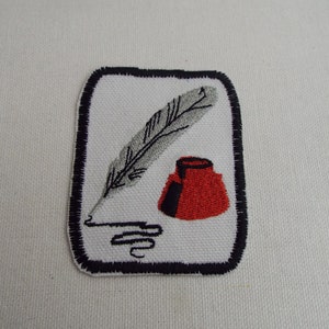 May include: A white embroidered patch with a black border. The patch features a gray feather quill and a red inkwell.