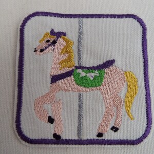 May include: Embroidered patch of a pink carousel horse with a green saddle and a purple mane and tail. The horse is on a silver pole against a white background. The patch is surrounded by a purple border.