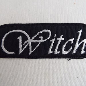 以下が含まれることがあります： 黒い刺繍パッチに銀色の糸で「Witch」と刺繍されています