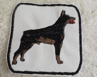 May include: Embroidered patch of a black and brown Doberman Pinscher dog with a red tongue sticking out. The patch is square and has a black border.