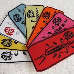 Op de afbeelding: Een set van tien geborduurde strijkpatches met een zwart dolk dat een rode roos met groene bladeren doorboort. De patches zijn verkrijgbaar in tien verschillende kleuren: wit, geel, oranje, roze, rood, blauw, groen, paars, bruin en zwart.