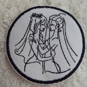 May include: Black and white embroidered patch of two people in wedding attire, with veils and flowers in their hair, embracing.