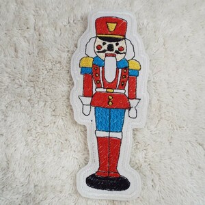 May include: A red and blue embroidered nutcracker with a yellow hat and a white background. The nutcracker has a black mustache and a red mouth.