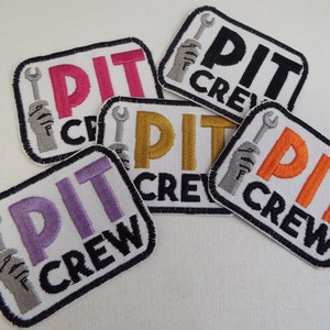 Peut inclure: Cinq patchs brodés thermocollants avec le texte "PIT CREW" en différentes couleurs. Les patchs présentent une clé à molette et une main tenant la clé à molette.