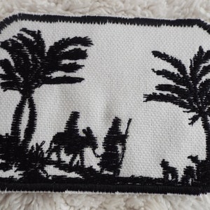 May include: Black and white embroidered patch depicting three figures walking with a camel in a desert landscape with palm trees.