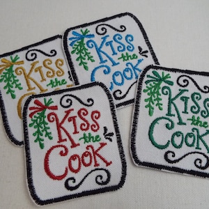 May include: Four embroidered iron-on patches with a white background and black border. Each patch features a green sprig of mistletoe and the text "Kiss the Cook" in different colors: gold, red, blue, and green.