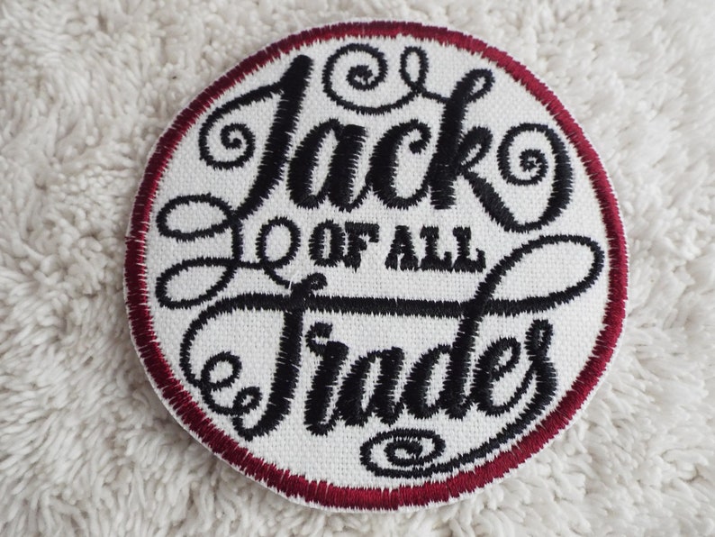 Op de afbeelding: Een witte geborduurde patch met een rode rand. De patch heeft de tekst "Jack of all Trades" in een zwart cursief lettertype.