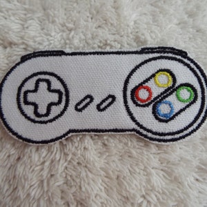 Op de afbeelding: Witte geborduurde patch van een Super Nintendo-controller met zwarte contouren en gekleurde knoppen.