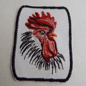 May include: Embroidered patch featuring a detailed rooster head. The rooster has a vibrant red comb and wattle, with black and white feathers. The patch is rectangular with rounded corners and a black border.