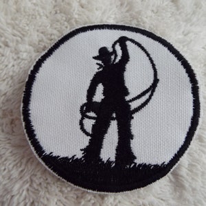 Puede incluir: Parche bordado en blanco y negro de la silueta de un vaquero sosteniendo un lazo. El parche es circular y tiene un fondo blanco con un borde negro.