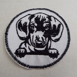 May include: Black and white embroidered patch of a dachshund puppy with its head peeking over a surface. The patch is circular and has a black stitched border.