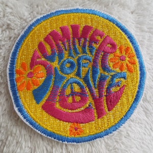 May include: A round, yellow embroidered patch with a blue and pink design. The text "Summer of Love" is embroidered in a stylised font with flowers surrounding the text.