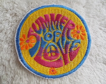 May include: A round, yellow embroidered patch with a blue and pink design. The text "Summer of Love" is embroidered in a stylized font with flowers surrounding the text.