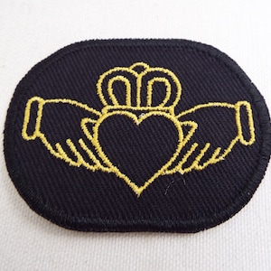 Könnte beinhalten: Schwarzer ovaler Aufnäher mit goldgesticktem Claddagh-Design. Das Claddagh-Symbol mit Herz, Krone und Händen steht für Liebe, Loyalität und Freundschaft. Der Aufnäher ist aus einem strukturierten Stoff gefertigt.