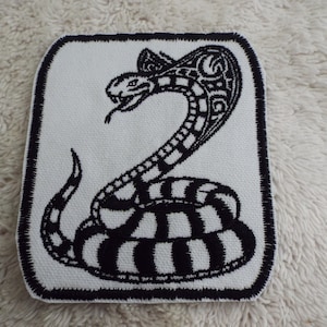 May include: A square, white patch with a black embroidered cobra design. The cobra is depicted with its hood raised and tongue extended, with a detailed pattern. The patch has a black border.
