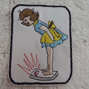 May include: Embroidered patch of a cartoon girl in a yellow and blue dress standing on a scale with a red needle pointing up. The scale is labeled with the number "1".