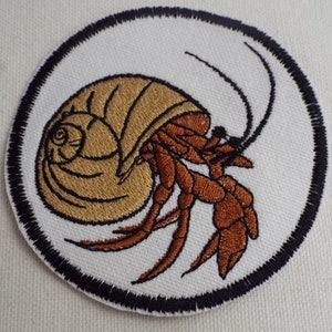 May include: Embroidered patch of a brown hermit crab in a shell, with black outlines, on a white background with a black circle border.