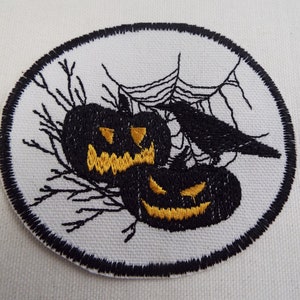 May include: A white embroidered patch with a black circle border. Inside the circle are two black jack-o'-lantern faces with yellow eyes and mouths. A black crow is perched on a web above the pumpkins.