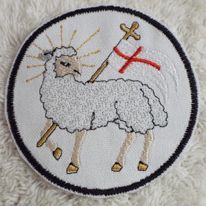 May include: Embroidered patch featuring a white lamb carrying a white flag with a red cross. The lamb is surrounded by a golden halo and the patch has a black border.