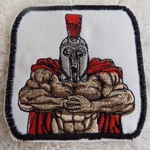 May include: Embroidered patch depicting a muscular, armoured warrior with a red cape and helmet. The warrior has a serious expression and is standing with arms crossed.