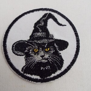 May include: Black embroidered iron-on patch of a cat wearing a witch's hat with a pointed brim. The cat has yellow eyes and a white face.