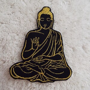 May include: Gold embroidered Buddha patch on a black fabric background. The Buddha is sitting in a meditative pose with hands in his lap.
