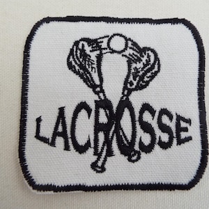 Puede incluir: Un parche bordado blanco con un borde negro. El parche presenta un gráfico negro de dos palos de lacrosse cruzados y la palabra "LACROSSE" en letras negras.