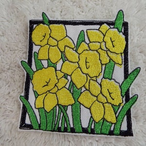 May include: An embroidered patch featuring a cluster of yellow daffodils with green leaves, framed in a black border.