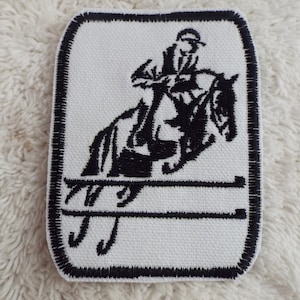 May include: Black and white embroidered patch of a rider jumping over a fence on a horse. The patch has a black border.