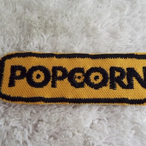 Puede incluir: Un parche bordado amarillo y negro con la palabra "POPCORN" en letras negras.