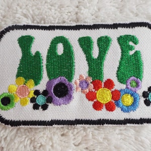Può includere: Un cerotto di stoffa bianco con bordo nero, che presenta la parola "LOVE" in lettere ricamate verdi. Le lettere sono circondate da fiori ricamati colorati.