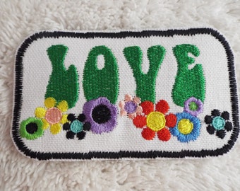 May include: A white fabric patch with a black border, featuring the word "LOVE" in green embroidered letters. The letters are surrounded by colorful embroidered flowers.