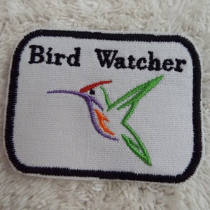 Könnte beinhalten: Weißer gestickter Aufnäher mit schwarzem Rand. Der Aufnäher zeigt den Text "Bird Watcher" und einen bunten Kolibri mit grünen, violetten, orangefarbenen und roten Akzenten.