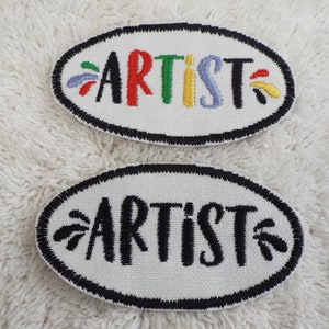 Puede incluir: Dos parches ovalados blancos con costuras negras alrededor de los bordes. El parche superior tiene la palabra "ARTIST" bordada en hilo de colores. El parche inferior tiene la palabra "ARTIST" bordada en hilo negro.