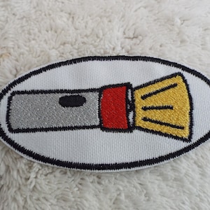 May include: A white oval embroidered patch with a black outline. The patch features a gray and red flashlight with a yellow beam.