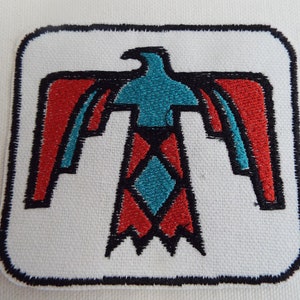 May include: A white square patch with a black outline, featuring a stylised red, blue, and black eagle design. The eagle is facing forward with its wings spread out.