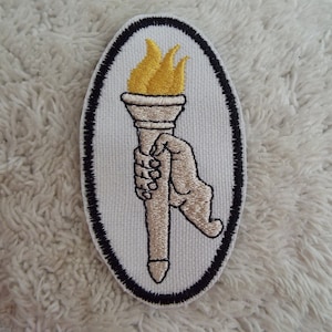 May include: A white oval patch with a black border, featuring an embroidered image of a hand holding a torch with a yellow flame.
