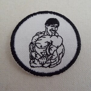 May include: A round, white embroidered patch featuring a muscular man flexing. The black outline of the man and the black border around the patch create a bold contrast. The patch is likely for clothing or accessories.