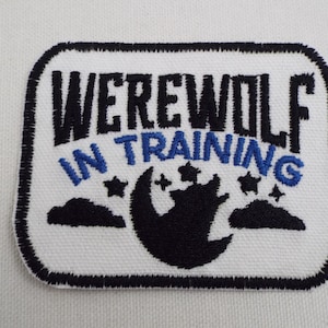 Könnte beinhalten: Weißer gestickter Aufnäher mit schwarzen und blauen Nähten. Der Aufnäher zeigt den Text "WEREWOLF IN TRAINING" mit einer schwarzen Silhouette eines Wolfes, der zu einem Halbmond mit Sternen heult.