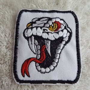 Puede incluir: Parche blanco bordado con una cabeza de serpiente negra y blanca con una lengua roja y ojos amarillos. La serpiente está en un marco cuadrado con un borde negro.