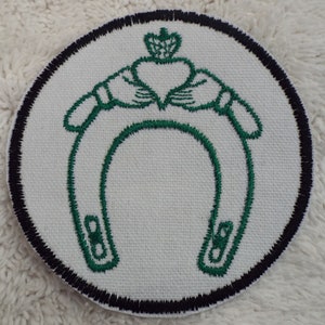 Puede incluir: Un parche redondo blanco con un borde negro. El parche presenta una herradura bordada en verde con un diseño de nudo celta encima.