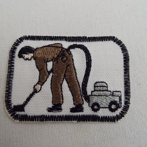 May include: A black and white embroidered patch depicting a person using a vacuum cleaner. The person is wearing brown pants and a brown shirt. The vacuum cleaner is silver and has a black hose.