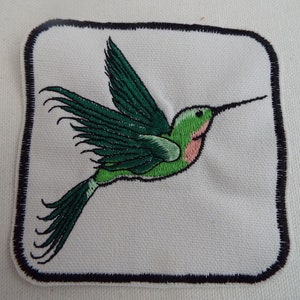 May include: A green and pink embroidered hummingbird patch with a black outline, on a white background.