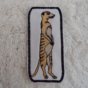 May include: A brown and white embroidered patch of a meerkat standing on its hind legs. The meerkat is outlined in black and the patch has a black border.