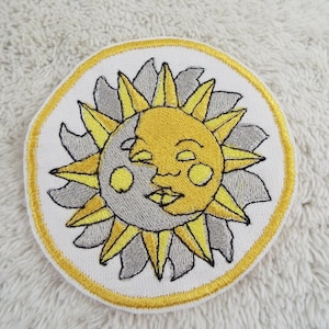 May include: A yellow and gray embroidered patch with a sun and moon design. The sun has a smiling face and the moon has a crescent shape.