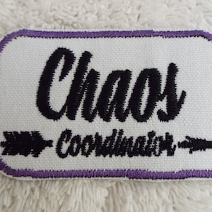 May include: White embroidered patch with purple stitching. The patch says "Chaos Coordinator" with an arrow pointing to the right.