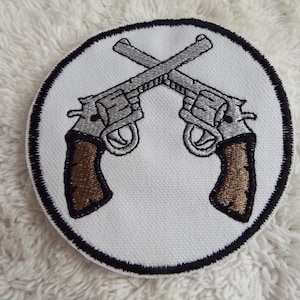 May include: A white embroidered patch with a black border, featuring two silver pistols crossed over each other with brown handles.